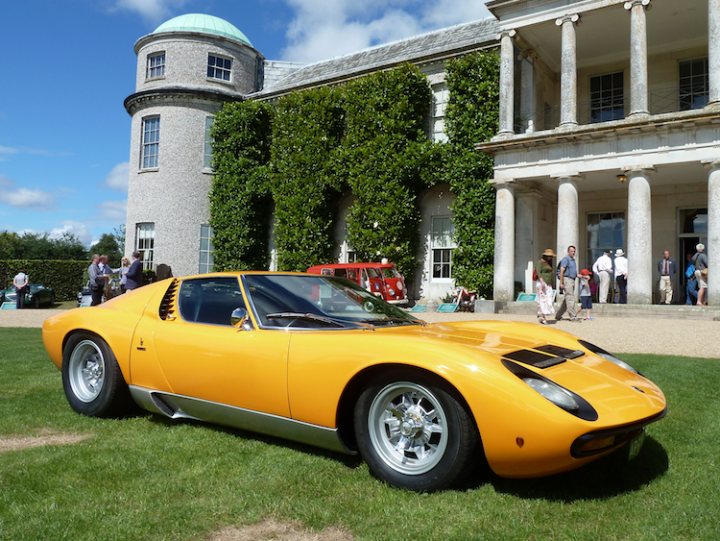 GRRC Open Day - 11 August - Page 1 - Goodwood Events - PistonHeads - The image features a vibrant yellow sports car parked on a well-maintained lawn in front of a large, historic building that boasts arched windows. A small group of people is scattered around the scene, with some standing closer to the car and others closer to the building. There are several other vehicles in the background, as well as a solitary figure on the right edge of the image. The building's grandeur, coupled with the classic design of the sports car, suggests a setting of stately elegance and leisurely activity.