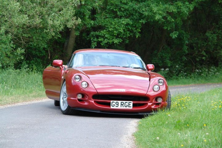 Just a little vid of my cerbera - Page 1 - Cerbera - PistonHeads - The image captures a dynamic scene on a winding road. A bright red sports car, bearing the license plate G9 RGE, is driving away from the camera. The car is sleek and powerful-looking, suggesting it's full of speed and excitement. The surroundings hint at a rural setting, with trees and foliage lining the road. The angle of the image gives a sense of motion and speed, as if the viewer is watching the car from behind and going along for the ride. The vibrant red of the car stands out against the greenery, adding to the dramatic feel of the scene.