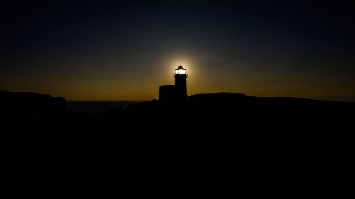 Drone Pics - Page 40 - Photography & Video - PistonHeads - This image features a lighthouse bathed in the warm light of the evening sun. The lighthouse is straddled between two rocky hilltops, creating a silhouette against the night sky. The dark blue of the night sky is set on a gradient where only the very top hints of a still visible sun. The sun casts a perfect semicircle illuminating the lighthouse and its surrounding cliffs, creating a picturesque scene often associated with coastal landscapes.