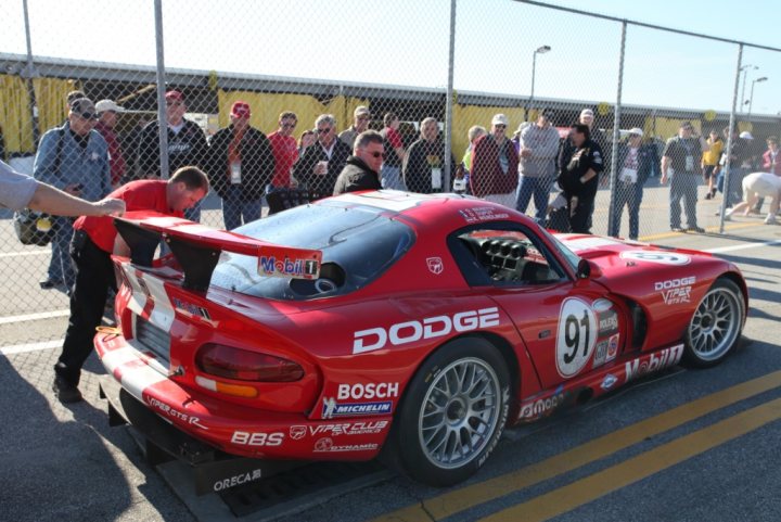 Daytona 24hr tour  - Page 1 - Vipers - PistonHeads - The image showcases a vibrant scene at a car show, with a striking red and white Dodge Viper GTS-R parked on a yellow boundary line. The car is the center of attention, with a group of people gathering around it, standing behind a fence, and observing it with interest. The Viper features a distinctive hood with a large scoop, indicating its high-performance capabilities. The license plate reveals it as a California-registered vehicle, adding a touch of mystery to the car's origin. The event appears to be an outdoor show, possibly near a racetrack, given the presence of tents and the excitement in the air.