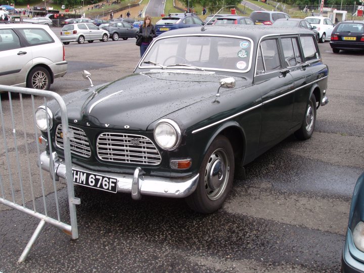 COOL CLASSIC CAR SPOTTERS POST!!! - Page 175 - Classic Cars and Yesterday's Heroes - PistonHeads - The image depicts a parking lot where a black Volvo station wagon is the main focus. The car is positioned adjacent to a temporary metal barrier, suggesting a restricted area or event. The photograph captures a clear day, and in the background, other cars can be seen, indicating a public place, possibly a dealership or parking garage. A solitary pedestrian is also present in the scene. The image appears to have been taken by an enthusiast or onlooker, rather than a professional photographer, which lends a candid and informal quality to the scene.