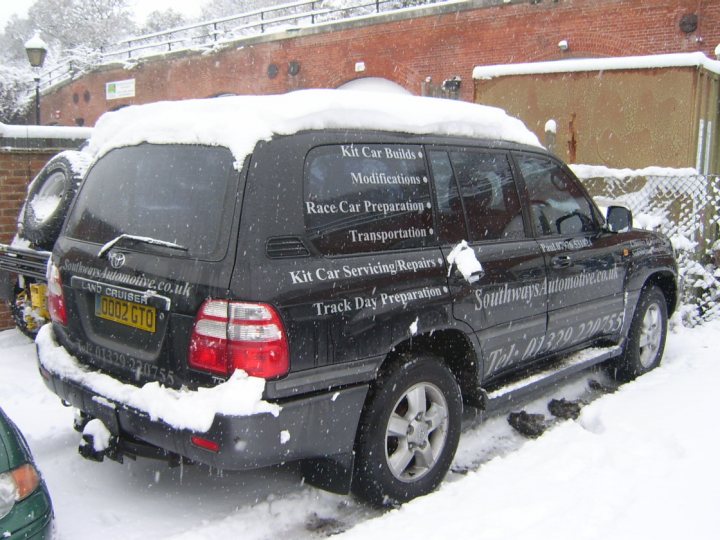 Which 4x4(s) have you got? - Page 1 - Off Road - PistonHeads - The image shows a black SUV parked on the side of a street, which is covered in snow. The vehicle is loaded with a significant amount of snow on its roof, indicating recent snowfall. Additionally, there are signs prominently displayed on the rear windshield of the SUV, suggesting that this vehicle might be used for commercial or service purposes, possibly offering services such as car building modifications and preparation, track day support, and track preparation services. These signs, along with the snow-covered vehicle, give a sense of a location possibly in the north during the winter season.