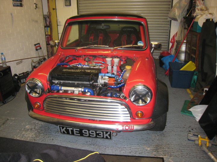 A red truck parked in front of a building - Pistonheads - This image depicts a vivid red vintage Mini Cooper car, parked within a storage space with a rolled-up carpet on the floor. The car's hood is propped open, revealing a well-maintained engine below. The engine bay and the structure are organized with items such as a fire extinguisher, paint cans, and various tools. The car's license plate reads "KTE993K," and on the side, there is a "Tow" sign that reads "1 TOW." This scene suggests the car is either in the process of receiving maintenance or has recently been completed.