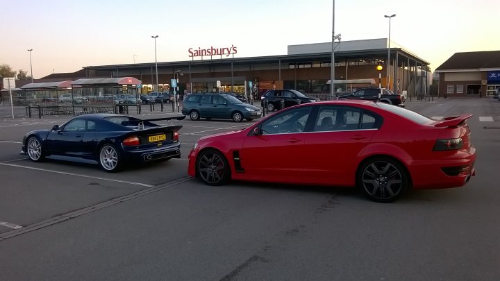 A car that is sitting in the street - Pistonheads - In the parking lot, lined with spaces, two cars stand out: a red sports car and a blue race car. The red sports car is parked close to the blue race car, creating a striking contrast with its vibrant red color. 

A handful of other cars are parked in the lot, but these two are the most eye-catching. In the background, a Sainsbury's supermarket stands, its red facade matching the color of the sports car. 

The cars are parked on the left side of the picture, close to each other, suggesting that they belong to the same person or are at least part of the same group.