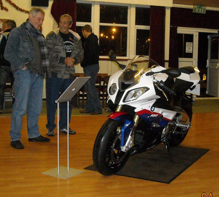 Gtg Plymouth Curry Pistonheads Xmas - The image showcases a gathering of men around a motorcycle display. The motorcycle, with its white and black design, is prominently placed in the center of the frame, attracting the attention of the spectators. Two men are standing close to the bike, while in the background, other men are observing the display. The scene is set in an indoor facility, likely a workshop or exhibition hall, characterized by a hardwood floor and an exit sign visible in the top right corner. The atmosphere suggests an interest in motorcycling, possibly a show, sale, or gathering of enthusiasts.