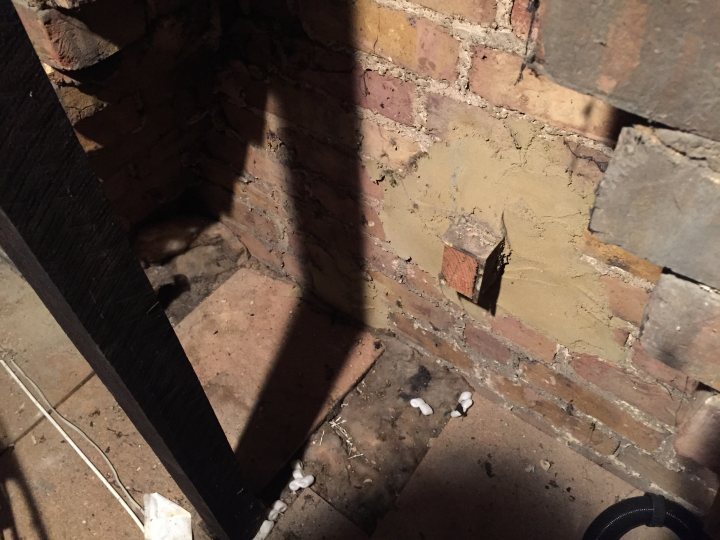 Falling debris inside bedroom wall - Page 1 - Homes, Gardens and DIY - PistonHeads - The image presents a dark and abandoned setting. The interior view of a tall room reveals a rough and bare brick wall that requires repair. A shadowy figure can be seen crouched in the corner of the room, adding an eerie touch to the scene. The overall atmosphere suggests neglect and disuse.