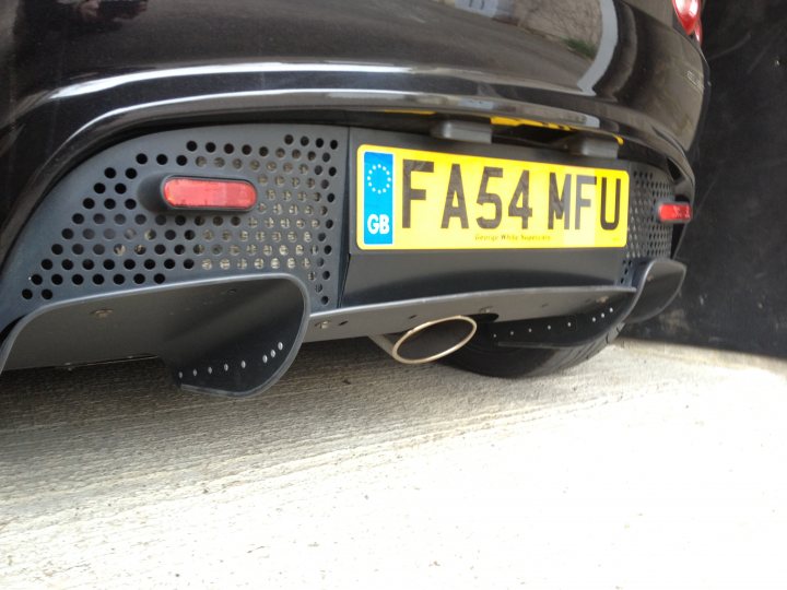 Slippery Slope... - Page 1 - Elise/Exige/Europa/340R - PistonHeads - The image shows the back of a black car with visible exhaust pipes. The registration plate at the rear of the car is yellow and reads "F54 MFU." The design of the exhaust pipe is black and appears to have small, circular holes. The reflection of the car can be seen on a surface, indicating that the car is outdoors. The number of channels in the report is low, suggesting that the photo might have been taken quickly or without much detail.