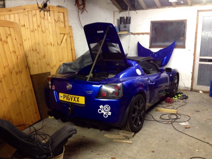 What did you do in the garage yesterday? - Page 5 - Chimaera - PistonHeads - The image shows a large, blue sports car situated on a garage floor. Its hood is hanging open, revealing the engine underneath. The car features thick protective striping on its sides, with the word "PIGVXX" prominently displayed, along with a logo that includes a drawing of a skull and crossbones. Additionally, there is a license plate that reads "P1GVXX" on the back. A wooden cabinet or toolbox is partially visible in the background. The ceiling of the garage suggests a rural setting due to the rafters and exposed brickwork. The lighting indicates dim, artificial light which creates a somewhat subdued atmosphere. The presence of tools and wires on the floor suggests ongoing maintenance or modification of the car.