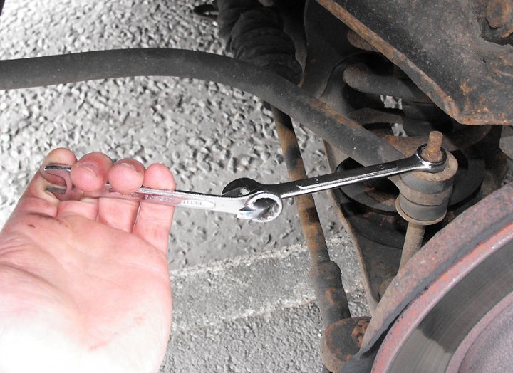 Help with stuck bolts - Page 1 - Home Mechanics - PistonHeads - In the background, a human hand is using a wrench to turn a nut. The wrench is black and silver, suggesting it might be silicone-coated to reduce slip. The nut being turned is attached to a rusty pipe. The rust suggests that this could be an old vehicle or equipment that has been in use for a considerable amount of time. The surface appears to be made of some kind of concrete or cement, adding to the overall worn and weathered look of the scene.