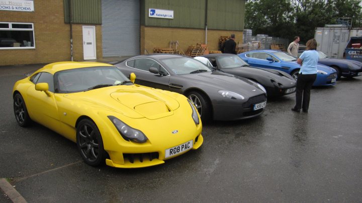 Pistonheads - The image captures a collection of four sports cars neatly parked in a lot adjacent to a brick building. These cars are all facing the same direction, exhibiting a sleek and shiny appearance. An interested individual can be seen closely examining the blue car in the middle of the row. The scene conveys a sense of excitement and admiration for these high-performance vehicles.