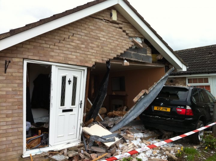 Pistonheads - The image captures a scene of domestic turmoil, with a black car and a red SUV having crashed into a house. The front door of the house is swung open, revealing a white doorframe backed by the interior wall. Scattered debris and an upturned section of the house's roof suggests structural damage. The cars are positioned as if an attempt to drive into or out of the home was made, with the front of the black car partially penetrating its interior. A red and white tape barrier is taped across the doorway, adding a dash of color to the scene.