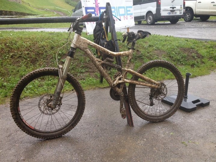 The "Photos From Today's Ride" thread... - Page 150 - Pedal Powered - PistonHeads - The image shows a mountain bike, prominently displayed in the foreground, leaning on a kickstand. The bicycle appears dirty, suggesting it has been used off-road or is awaiting cleaning. Behind the bike, there is a white van and a black vehicle partially obscured. The ground is wet, hinting at recent rain, and the surrounding area includes a grass-covered ground and a network of pavement strips, suggesting a rural or semi-rural setting.