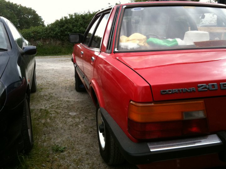 Cortina Ford Pistonheads - The image shows a red car parked on a patch of grass alongside a road. The car is angled to the viewer, with the rear side visible, and has a shiny texture. In the background, a green bush is visible, providing a contrasting backdrop to the vehicle. Another car is also partially visible in the foreground, indicating that the red car is not alone in the space. The overall scene suggests a casual, unhurried setting, possibly a residential area or a park with paved paths.