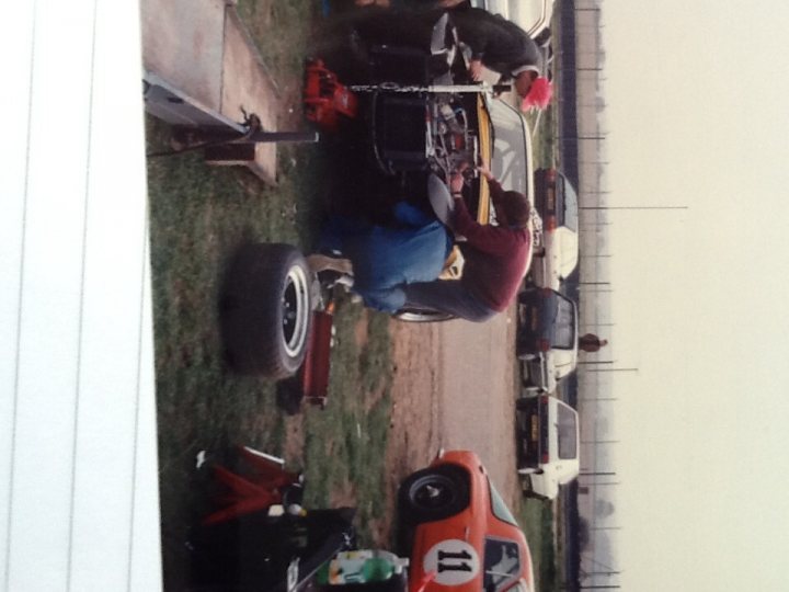 Early TVR Pictures - Page 10 - Classics - PistonHeads - The image depicts an outdoor vehicle repair scene with multiple cars parked on the grass, positioned in front of a red and white car. Several individuals, wearing clothing with different two-digit numbers, are working on the automotive repairs. There's a focus on a vehicle with an open trunk, which appears to be the primary object of attention. The setting appears to be an informal, temporary repair space, as suggested by the presence of a tool chest and the casual attire of the workers. The photo has a yellowish tone and gives a candid impression of mechanics at work.