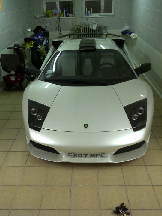 Pistonheads - The image shows a white vintage style sports car indoors, parked close to a blue motorcycle. The car has a distinctive headlight design and a black grille. The interior of this vehicle is not visible. The setting appears to be a garage or indoor repair shop, with a whiteboard against a wall in the background, and various items like parts or tools scattered around. The overall atmosphere suggests a space dedicated to automotive enthusiasts or mechanics due to its purposeful environment.