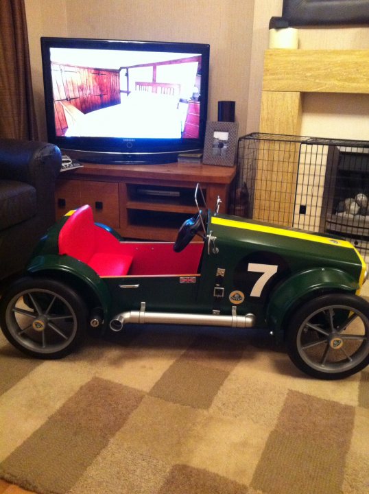 My 2yr old granddaughters new Lotus! - Page 1 - Elise/Exige/Europa/340R - PistonHeads - The image shows a vibrant green and yellow toy car parked in a living room. The car has the number 7 visible and appears to be a pedal car, given its design and small size. The interior of the room features an entertainment unit with a television displaying a blurry sports event, possibly a car race given the context. The room itself is furnished with a couch partially visible in the bottom left corner and a basketweave-style carpet underfoot. The mood of the image seems to evoke a sense of nostalgia and play.