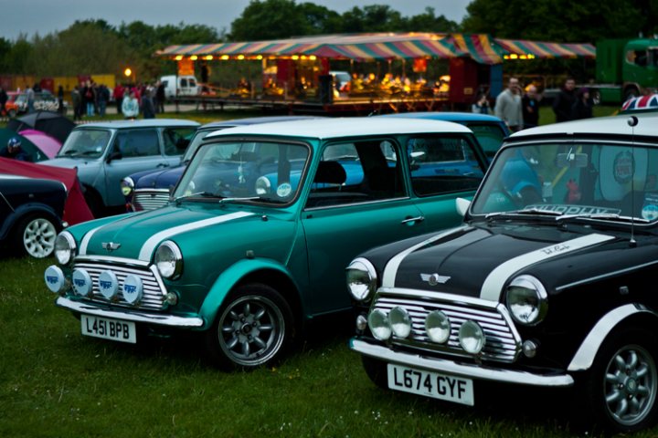 London to Brighton  - Page 1 - Classic Minis - PistonHeads - The image depicts a vibrant scene at an outdoor event. A classic turquoise Mini Minor stands out in the center-left, while a sleek black car with white stripes is positioned towards the right. Behind these cars, the event tent with its colorful roof is visible, providing a lively backdrop. Further to the right, under a green canopy, several passengers are standing, adding to the bustling atmosphere of the event. The lighting suggests it could be either morning or evening, given the soft illumination across the scene. The grassy area and the variety of stands suggest an outdoor, possibly festive event.