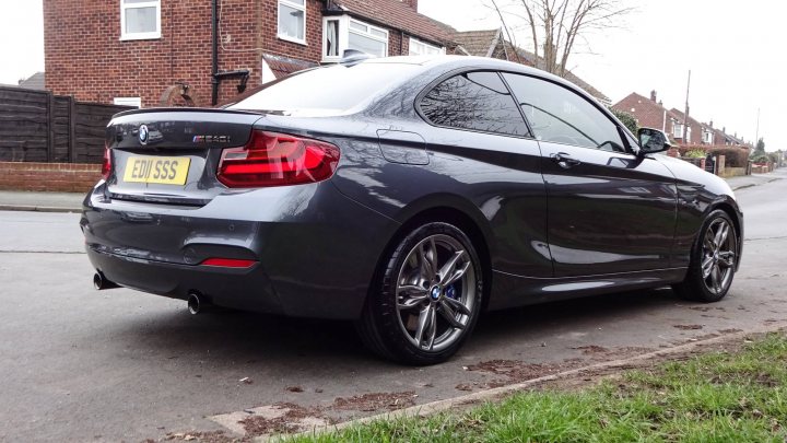 BMW M240I - Page 1 - Readers' Cars - PistonHeads - The image depicts a dark-colored BMW car parked on a residential street. The car is positioned parallel to the curb with a view from a pedestrian's perspective facing towards the rear left side of the vehicle. The car's tail lights and rear window are clearly visible, and the car is surrounded by a calm, suburban setting with a neatly manicured lawn and brick buildings in the background.