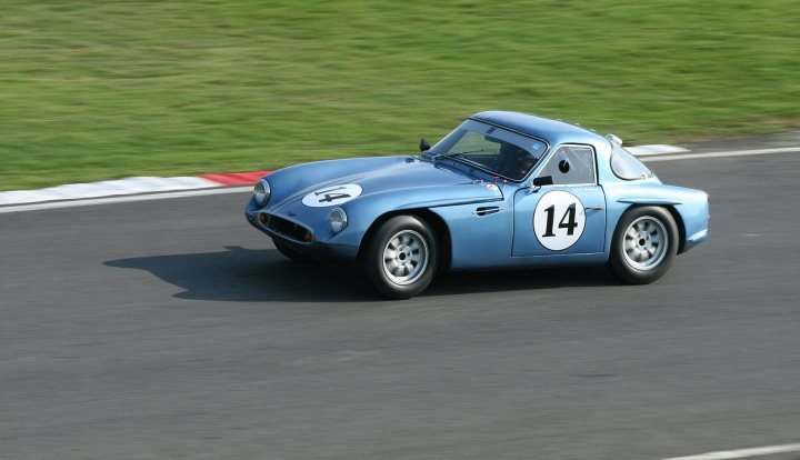 Early TVR Pictures - Page 126 - Classics - PistonHeads - The image shows a small, blue sports car with the number 14 prominently displayed on its side and hood. The car is sounding around a curve on a racetrack, giving the impression of speed. The surrounding green grass suggests the track may be outdoors. The photograph captures the car in motion from a side angle, which emphasizes its low and sleek design.