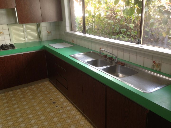 Pistonheads - The image displays a small kitchen with a counter area featuring a double basin sink. The sink appears to be made of stainless steel and is situated under a window with natural light coming through. The wall around the sink has a greenish tone, contrasting with the white-tiled area above the countertop. The kitchen cabinets are dark brown, and the floor is covered by linoleum tiles with an orange and cream pattern. The overall impression is that of a compact, functional space with a focus on the sink area.