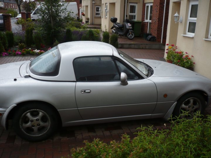 Booked Bodyshop Pistonheads - The image features a silver convertible car parked on a brick driveway. The car has a distinctive rear window design and a visible black side stripe. On the left side of the image, there is a building and a scooter parked on its stoop. The setting appears to be a residential area with potted plants visible. The car's roof appears to be down, suggesting it might be a convertible model. There are various potted plants and shrubbery around, adding to the residential feel of the image.