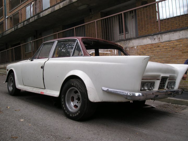 The image features a vintage white car parked on a street beside a tall, multi-story building. The car is a classic sedan with a distinctive elongated rear section. It has round, chrome detailing, including grilles, bumpers, and wheel accents. The red door of the car is open, suggesting recent use. The vehicle is equipped with a black roof rack on its roof, adding to its visual appeal and hinting at its potential function for transporting luggage or other items. The street appears to be quiet with no other vehicles in sight, which highlights the car as the primary subject of the image.