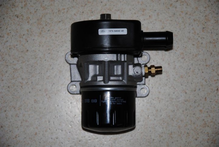 Pistonheads - The image shows a close-up of a black engine carburetor. It appears to be a mechanical part with a metallic finish, and there is a white label on the side with some text, likely providing information about the carburetor's model or brand. The top of the carburetor has a thin tube connected to it, which is probably the inlet of the fuel pathway. The background is a plain surface that looks like a floor or a countertop.