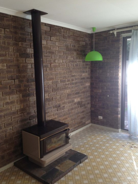 Pistonheads - The image shows a corner of a room where a modern wood-burning fireplace with a visible glass panel is integrated into a rustic brick wall. Hanging above the fireplace is a green pendant light. Adjacent to the fireplace on the right is a doorway leading to another room, and the floor is laid with small, square tiles. Sunlight appears to be entering the room through the doorway, illuminating the space and creating a warm, inviting atmosphere.