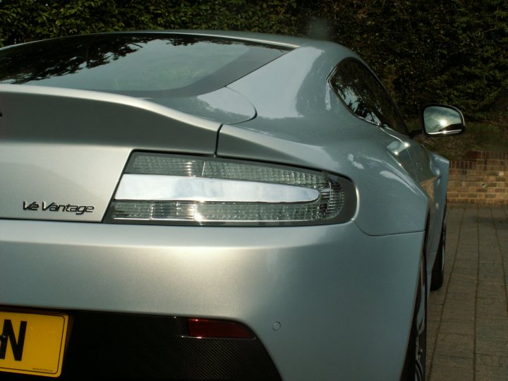 Dbs Pistonheads Vantagearrived - The image shows the rear side of a silver sports car, possibly a BMW or a similar make, with the back lit up. The car sports a sleek design, highlighted by a visible taillight and a license plate. The vehicle is parked on a brick surface, and there are trees and foliage in the background, suggesting an outdoor setting. The car's pristine condition implies it's well-maintained or has not been driven much.