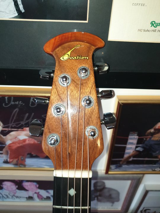 Lets look at our guitars thread. - Page 294 - Music - PistonHeads - The image shows a close-up of a guitar with a wooden finish. The guitar is attached to a wall and features multiple tuning pegs, indicating it's a six-stringed instrument. There are signatures on the front of the guitar, suggesting it may be autographed or signed by someone. In the background, there is a framed picture hanging on the wall with some text on it, but the details in the image are not clear enough to discern. The overall setting appears to be an indoor space, possibly a room dedicated to music or memorabilia.