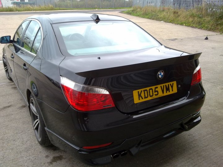 Pistonheads Kevin Bmw - The image is a photograph of a dark-colored BMW car, specifically the model name and year appear to be "kdos5vvy". The BMW model is reflected on the back window of the car. The vehicle is parked on concrete, facing away from the viewer, with the right side of the car visible in the foreground. The setting appears to be an outdoor parking area, potentially in a commercial or industrial district, based on the visible fence and some greenery in the background.