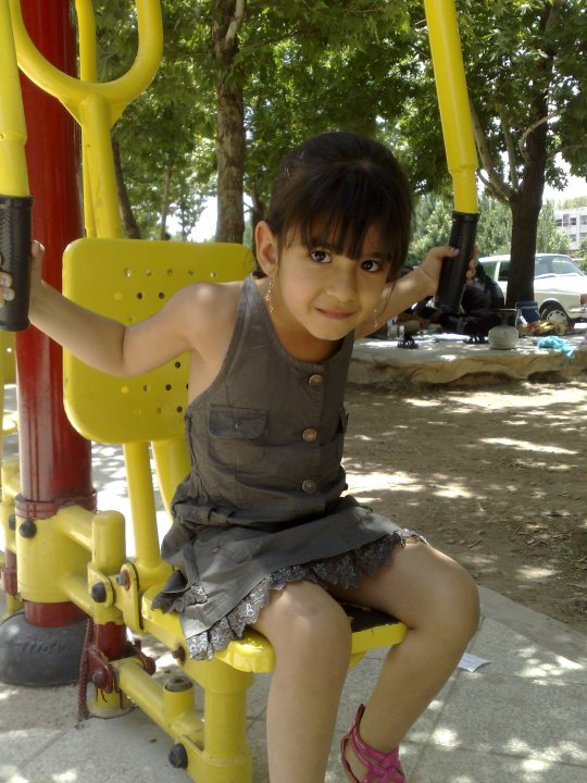 In the image, there's a young girl sitting on a yellow swing, smiling towards the camera. The swing is attached to a sturdy metal pole. Behind the girl, there's a park-like setting with a playground, trees, and a building in the distance. The joyful scene suggests it might be on a sunny day. The girl appears to be enjoying her day outside.