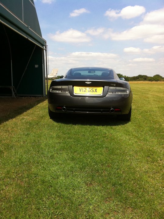 Pistonheads - This image shows a black sports car parked in an open field. The car's license plate reads "V12 GSX" and has a sleek design. The field is lush green, indicating it's likely spring or summer. In the background, against a blue sky dotted with clouds, there is a large green building with a grayish roof, possibly a barn or a new building in the field. The car's positioning and the clear sky suggest a calm and serene atmosphere.