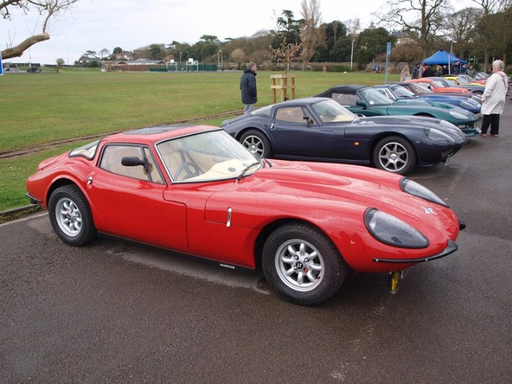 Marcos Display - Clevedon 14/04/2013  - Page 1 - South West - PistonHeads - In the image, a collection of sports cars is parked on a gravel lot, creating a vibrant display of colors. The red sports car is prominently visible in the foreground, with its glossy finish reflecting the surrounding environment. A few people can be seen walking around, admiring the cars. The event appears to be outdoors during the daytime, given the ample natural lighting. From the background, the silhouette of trees can be seen, adding a touch of nature to the scene.