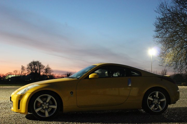 350Z GT4 - Ultra Yellow - Page 2 - Readers' Cars - PistonHeads - The image features a yellow sports car parked on a gravel surface. The car's sleek design and shiny exterior suggest it is well-maintained. The setting appears to be during the evening or early morning, given the lamplit in the background and the warm glow of the lights. The area around the car is devoid of other vehicles or significant activity. The sky is lightly adorned with clouds, and the tranquil atmosphere suggests a peaceful end or beginning of the day.