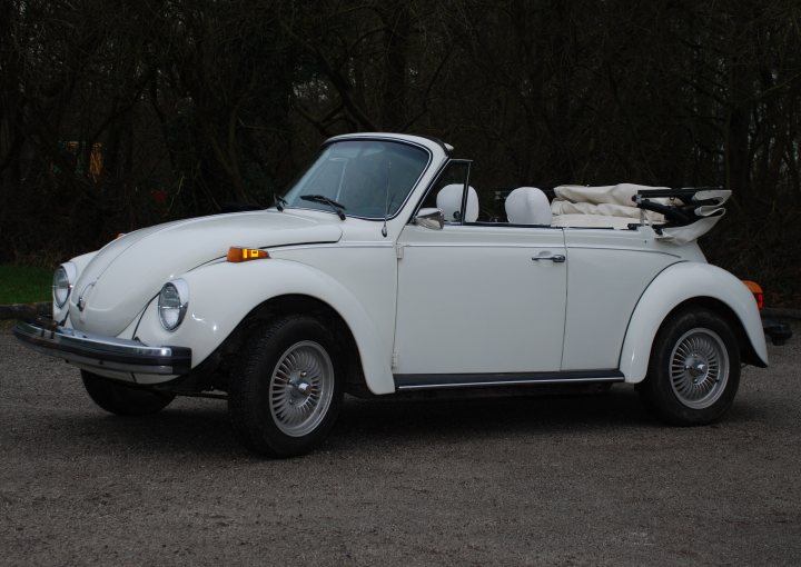 Need a Wedding Car For 11th May - Please Help - Page 1 - North West - PistonHeads - The image features a classic, vintage white Volkswagen Beetle convertible parked on a sparsely vegetated area that might be a dirt parking lot. This particular Beetle is characterized by its rounded white body, prominent spare tire mounted above the trunk, and its convertible roof, which is down in the photo. There's a licence plate at the front, and the car has a built-in luggage compartment at its rear, demonstrating the vehicle's design for practicality and style typical of the times when it was produced. The combination of the white color and the early model suggests this might be a rare or classic vehicle that retains its original form.