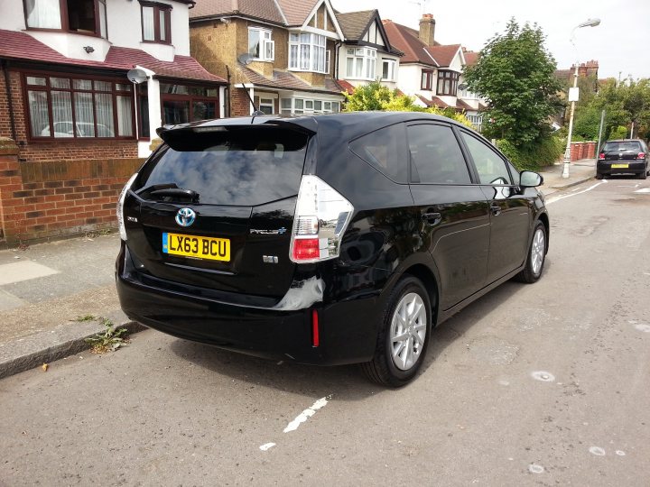 Toyota Prius Plus MPV - anyone got one? - Page 1 - Jap Chat - PistonHeads - The image presents a black Toyota hatchback positioned on a residential street. The car is parked perpendicular to the curb and is angled slightly towards the camera. The parked car is facing away from the viewer, showcasing the rear of the vehicle. To the left of the car, there's a brick building with a brown porch area. On the right, various green plants are visible, perhaps part of the garden or landscaping around the street. The scene captures a typical urban residential setting.