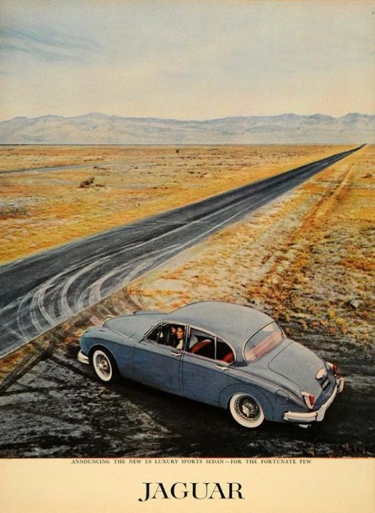 Pistonheads - This is a vintage advertisement featuring a blue Jaguar car parked on the side of a dirt road. The car has an old-fashioned design, suggesting it's from an earlier era. In the background, there is a desert landscape with a clear sky, reinforcing the sense of being in a remote location. The advertisement also includes text, which might contain branding or messaging related to the vehicle and its use in the context depicted.