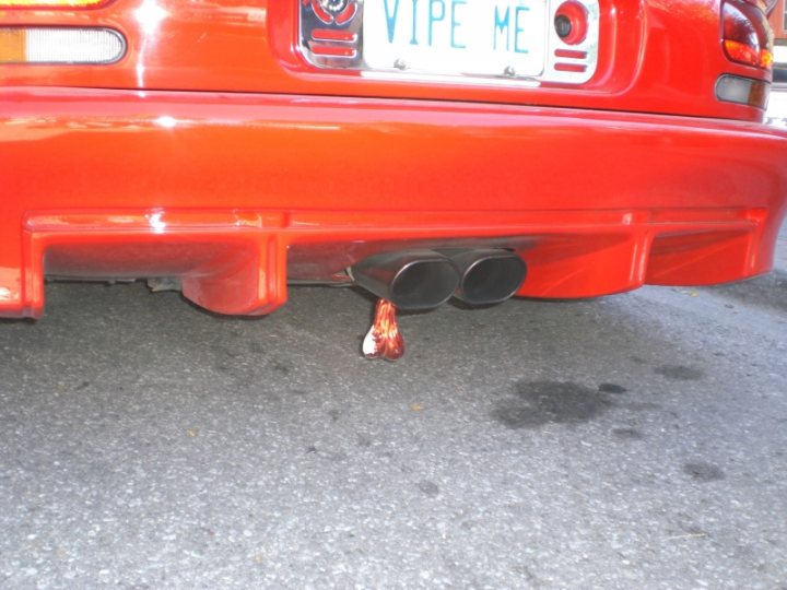 Tail Pistonheads Pipes - The image showcases the back bumper of a vibrant red car, highlighted by a distinctive black exhaust pipe. This car is registered in the state of Wisconsin, as indicated by the license plate that reads "VINGNES IS LVNGWITHD". A unique adding to the car's character is a red reflector, casually hanging from the exhaust pipe, adding a touch of humor or personalization to the vehicle.