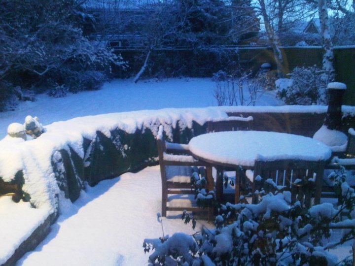 Installing Woodburner Pistonheads - The image captures a wintry outdoor setting where a light dusting of snow blankets the landscape. In the foreground, a snow-covered outdoor table and two benches are visible, suggesting a seating area. Lighting suggests that the photo may have been taken around dusk. In the background, another table with a bench and a couple of statues that are also dusted with snow. A fence runs along the yard, separating it from a wooded area visible in the distance. The scene conveys a calm and serene atmosphere typical of wintertime.