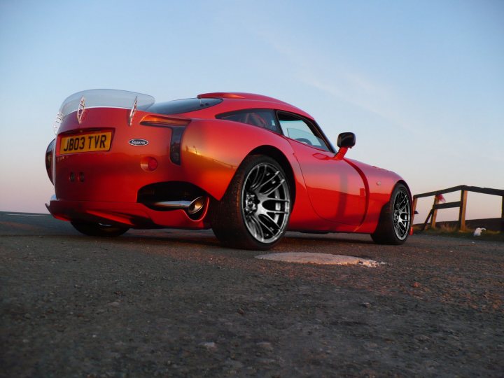 A red and blue car parked in a parking lot - Pistonheads - The image showcases a stylish, red sports car parked on a paved surface on a bright day. The car is equipped with a large rear spoiler to manage airflow, and its wheels are adorned with a silver and black color scheme. It has distinctive side vents and a concept-like design. The backdrop of pastel colors from the sky enhances the overall aesthetics of the image. The car seems to be alone, without any visible people or other objects in the immediate vicinity.