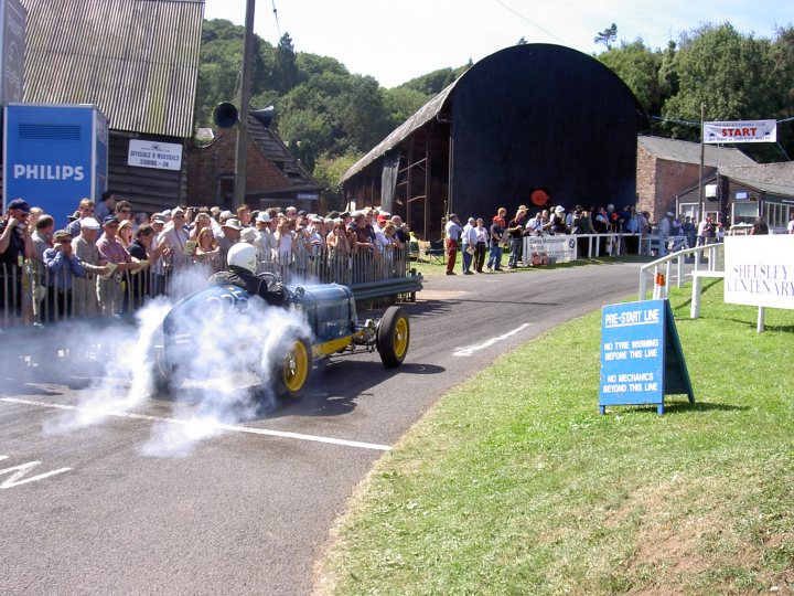 Meeting Classic Pistonheads - The image features a vintage smoke-blowing vehicle, possibly a steam tractor or a vintage firetruck, in the process of creating a cloud of smoke. The vehicle is driving down a road surrounded by a grassy area that looks like a race track, with spectators on both sides of the road. Some of the spectators are behind a barrier, and others are standing directly next to the road. To the right, there is a blue sign with white lettering mounted on a post. The setting appears to be an outdoor event or performance.