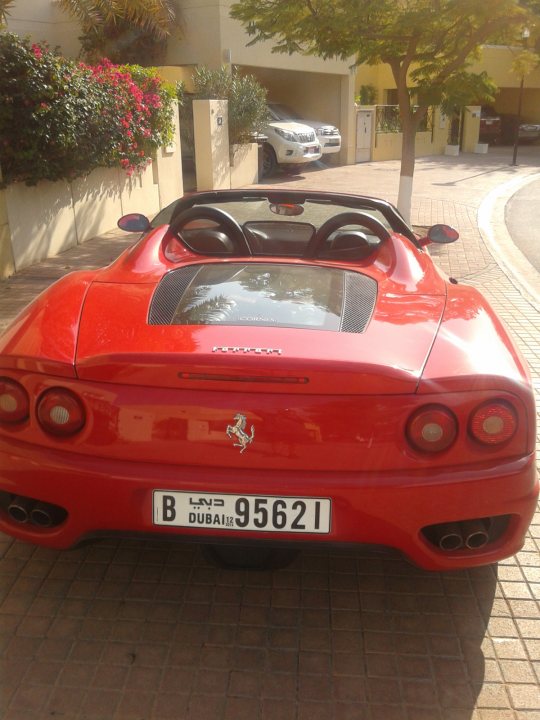 A red car is parked in a parking lot - Pistonheads - The image showcases a sleek red sports car parked on a brick-paved driveway in front of a garage. The car features a two-seater convertible design with a black roof that retracts, creating an open-top experience. On the rear of the car is the luxury brand's emblem, indicating it is a high-end vehicle. The license plate reads "B LND 95 621," which could suggest it's a luxury vehicle model with a distinctive set of identification numbers. In the background, there are other cars and buildings, indicating an urban or residential area.