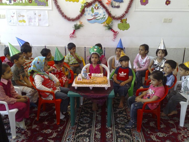 The image depicts a joyful birthday party. A group of children are gathered in a room with a festive atmosphere. They are seated in a formation around a table, some with colored party hats on their heads. A small birthday cake is placed on the table, with lit candles indicating that it's time for birthday greetings and candle-blowing. The room is decorated with children's crafts, adding to the lively and celebratory ambiance of the party.