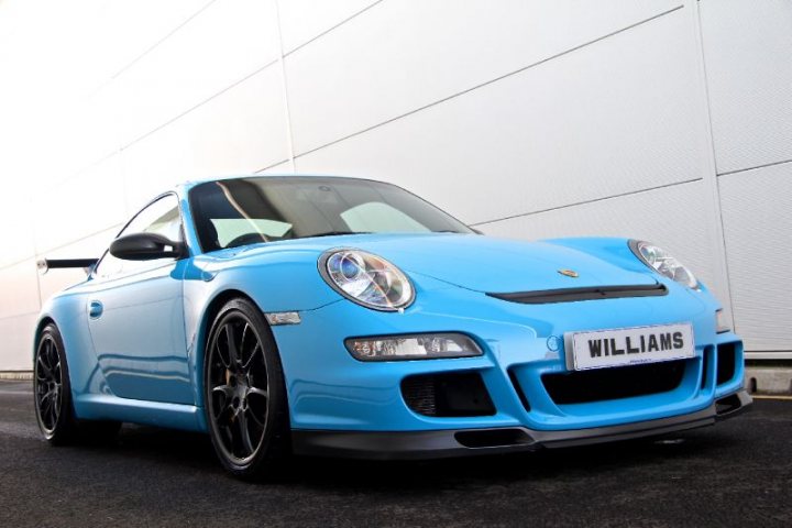 A blue car parked in a parking space - Pistonheads - The image features a vibrant blue Porsche sports car positioned at a slight angle, showing its side profile. The car is parked on a tarmac that is bordered by a fence and a metal structure, suggesting it's located in a secure parking area. The reflections on the car's shiny exterior indicate that the picture is either taken in the early morning or on a rainy day. The overall setting seems to be outdoors, given the exposed area surrounding the car.