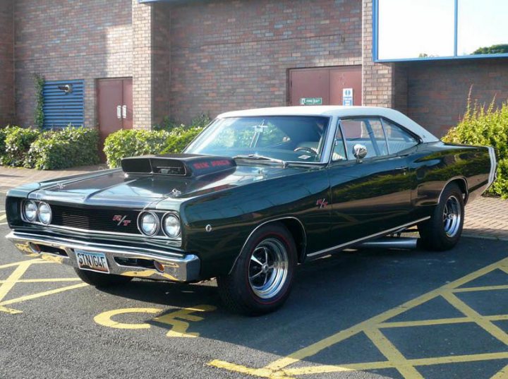 Pistonheads - The image shows a classic black car, which appears to be a muscle car based on its design, parked in a parking lot. The car features a prominent hood scoop and flared wheel arches, indicative of a performance vehicle from a previous era. It is positioned with two wheels visible in the frame, and the side includes distinctive emblems, likely on the doors, which are typical of iconic muscle cars. The setting is a paved parking lot with a marked parking space visible under the vehicle, and there is a structure with a blue sign in the background. There are no visible texts in the image to provide additional context.