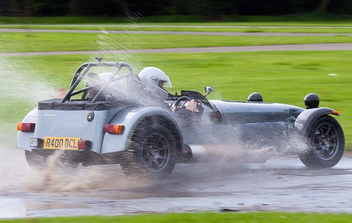 Pistonheads - The image captures a dynamic scene of a silver racecar driving on a wet track. The car is in action, its tires creating a spray of water as it accelerates. The driver, clad in a white helmet and visible through the windshield, is focused on the road ahead. The background reveals a grassy field, reinforcing the sense of a racing event in an open, outdoor setting.