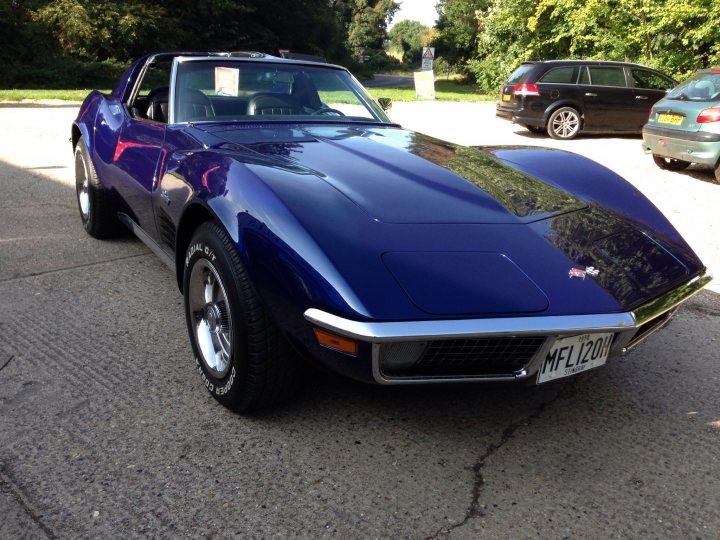 A car is parked in a parking lot - Pistonheads - The image shows a striking purple sports car parked on a tarmac, viewed from the front left side. The vehicle is sleek, with a long sleek hood and a short tail, indicative of a classic sports car design. The brake dust can be seen on the front wheel and markings visible in the concrete. In the background, there is another vehicle parked, partially obscured by the sports car, and some green vegetation, suggesting a park or a garden.