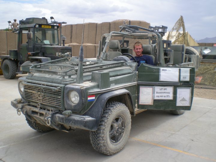 Difference Wagon Mercedes Pistonheads - The image depicts a scene featuring a gray military vehicle parked outdoors. Inside the vehicle, there's a person in the driver's seat, and a soldier is seen sitting in the hatch at the back of the vehicle. The vehicle displays dusty and weathered condition, suggesting it's been in use. In the background, there's a partial view of other military vehicles and a sandbagged wall, suggesting a Military base or secure area.