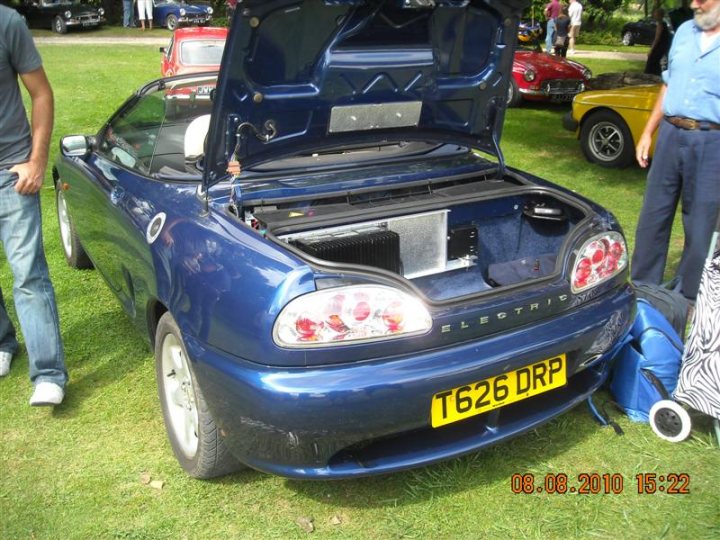 Pistonheads - The image displays a blue, two-door sports car in the process of being viewed or prepared. The hood of the car is open, revealing a partially disassembled trunk area. A man is peering into the back of the car, likely inspecting or organizing the contents. The car is parked in a grassy area, with other cars and individuals in the background, suggesting an outdoor gathering or event. Other objects, including a luggage bag and a clear plastic bag, can be seen in close proximity to the car, indicating recent activity around the vehicle. The timestamp in the corner of the image indicates it was taken in October 2010.